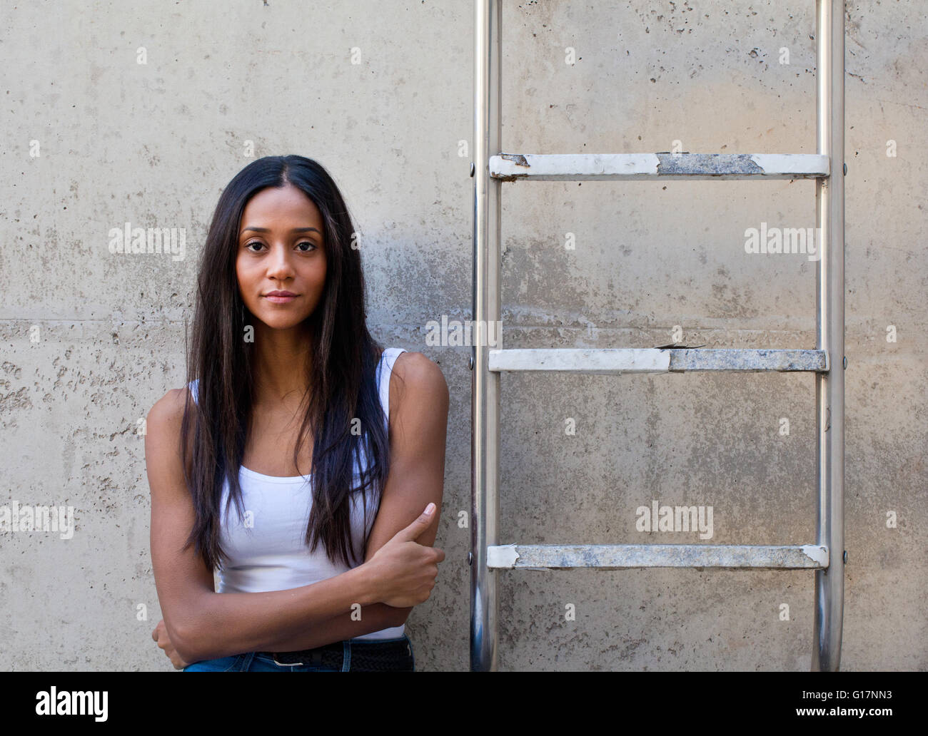 Woman leaning against concrete wall next to ladder, arms crossed looking at camera Stock Photo