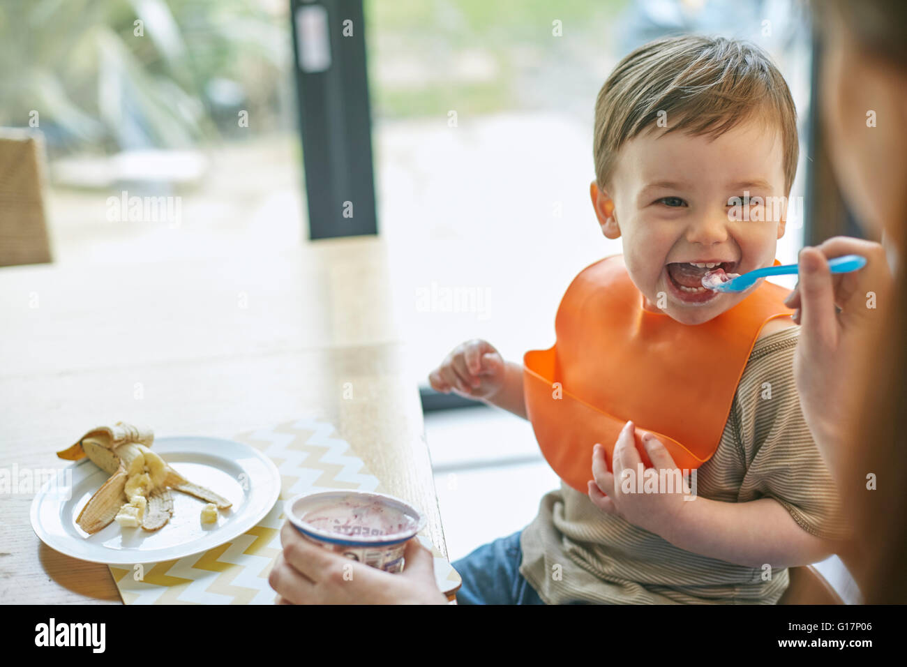 Happy baby boy sitting at table being fed yogurt by mother Stock Photo