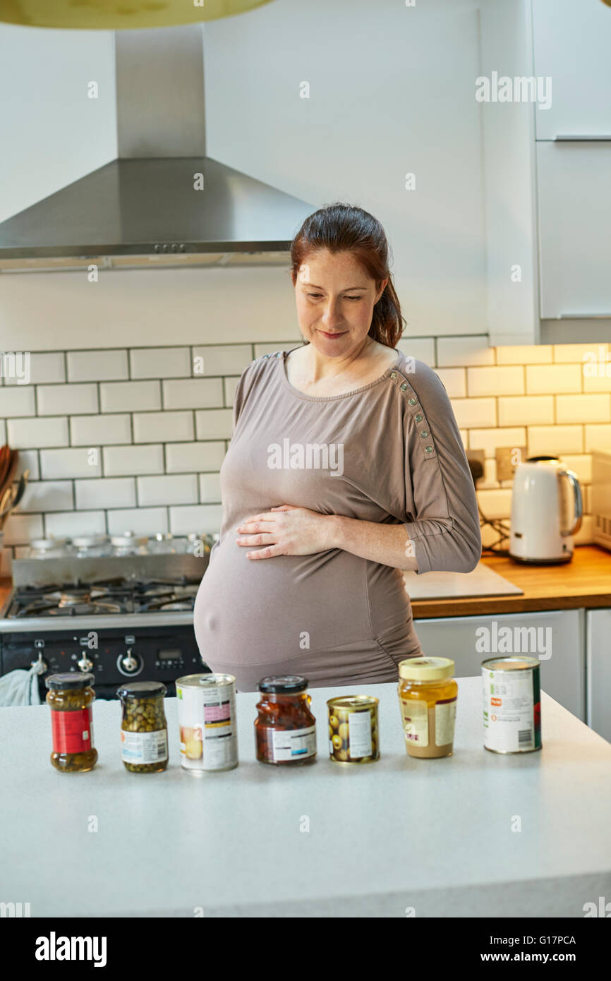Pregnant woman in kitchen looking at jars and tins of food on kitchen counter Stock Photo