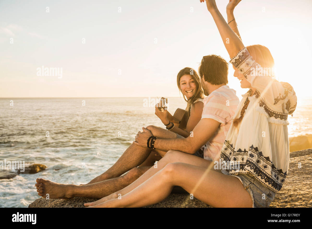 Three mid adults sitting on rocks playing acoustic guitar at sunset, Cape Town, South Africa Stock Photo