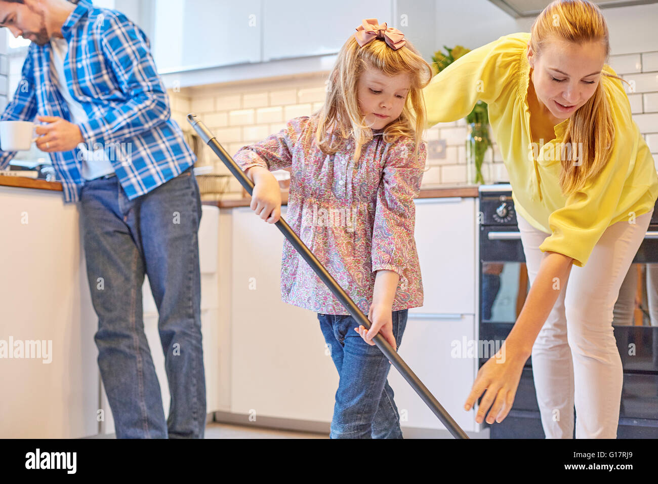 Mother and daughter mopping kitchen floor, father reading in background Stock Photo
