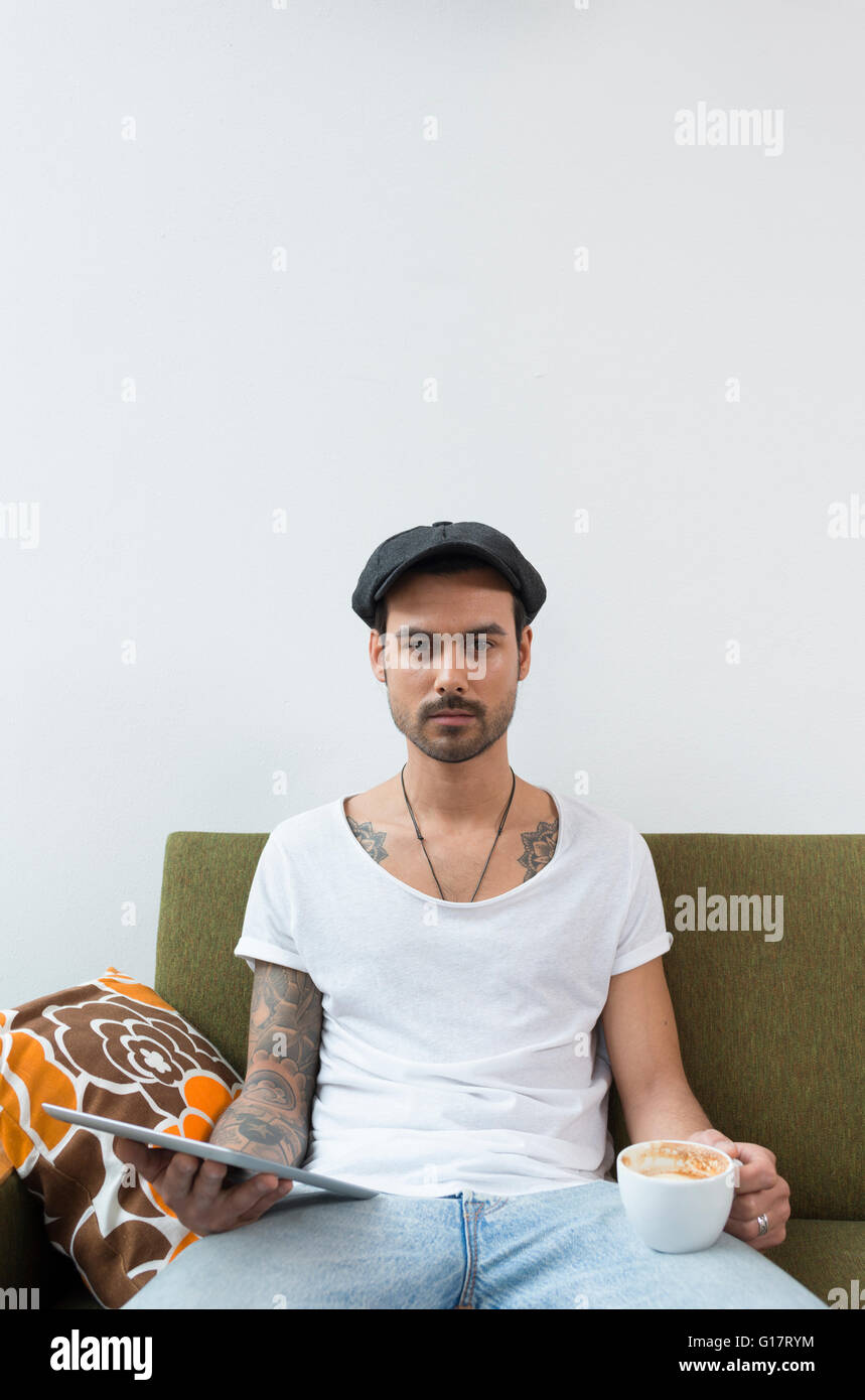 Portrait of young male hipster sitting on cafe sofa with coffee cup and digital tablet Stock Photo