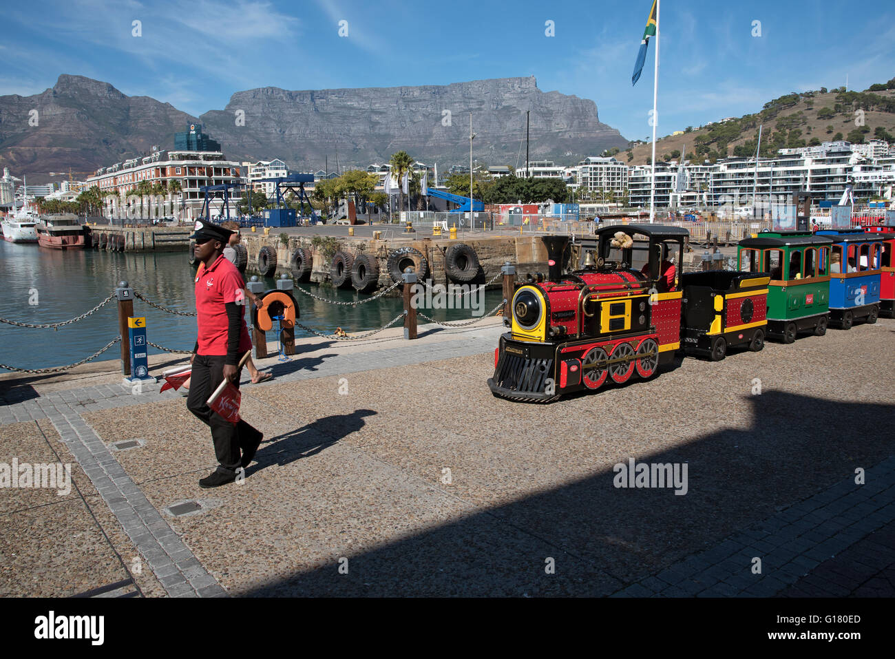 CAPE TOWN WATERFRONT SOUTH AFRICA  Man in uniform carrying a red flag with a children's train ride around the waterfront Stock Photo