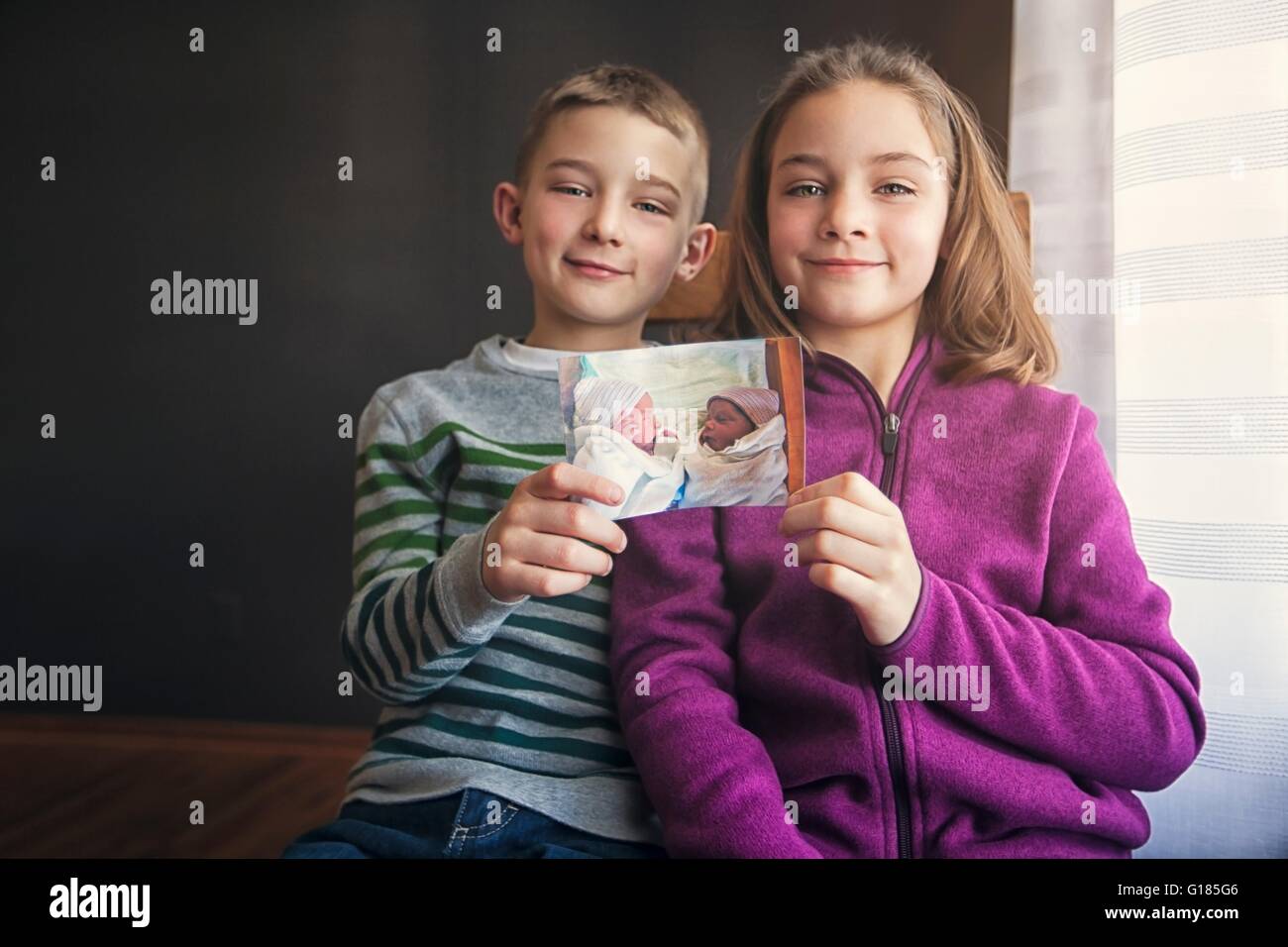 Portrait of boy and girl twins holding up photograph of themselves as babies Stock Photo