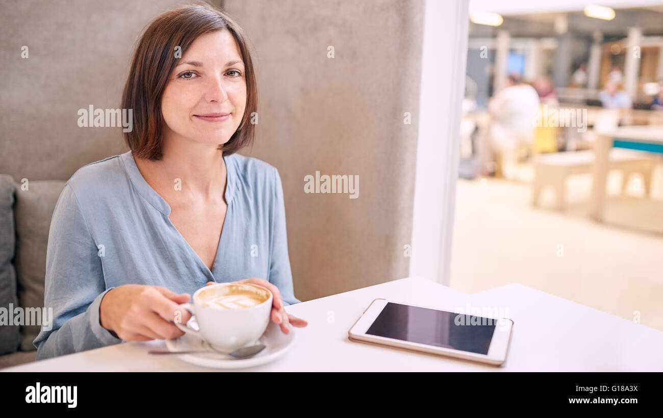 mature caucasian woman smiling at the camera while sitting in a well lit coffe shop with a cafe latte in her hands and her elect Stock Photo