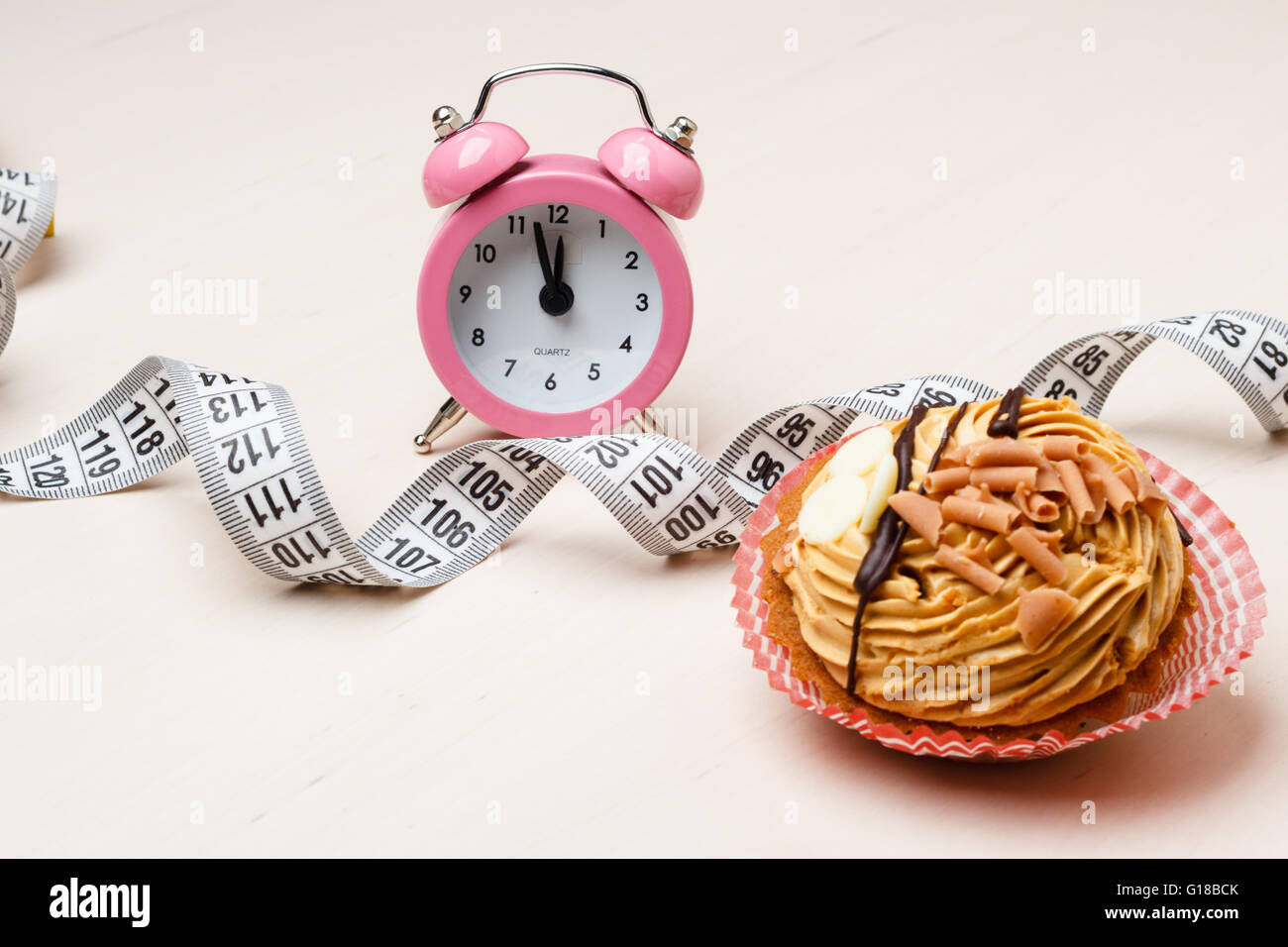 Gluttony and not eat junk sugar foods concept. Time for slimming. Cake cupcake measuring tape and alarm clock on kitchen table Stock Photo