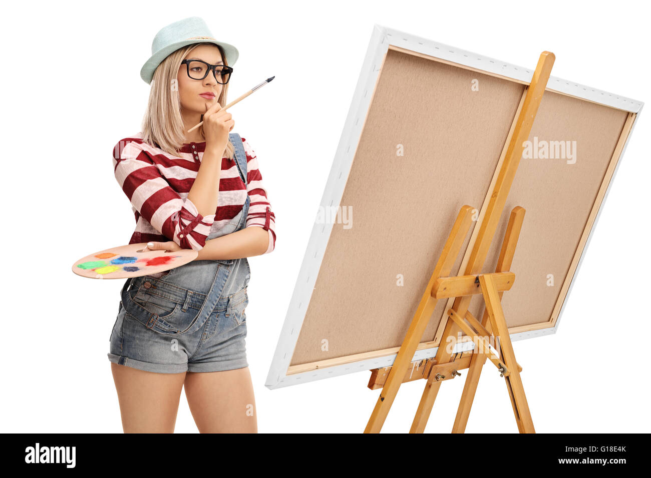 Female artist holding a paintbrush and a color palette and looking at a painting isolated on white background Stock Photo