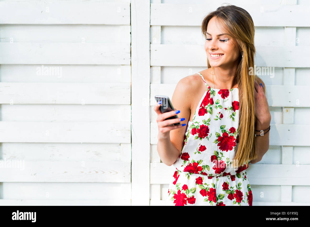 Beautiful young woman using phone while leaning against a white wall during a bright summer day Stock Photo