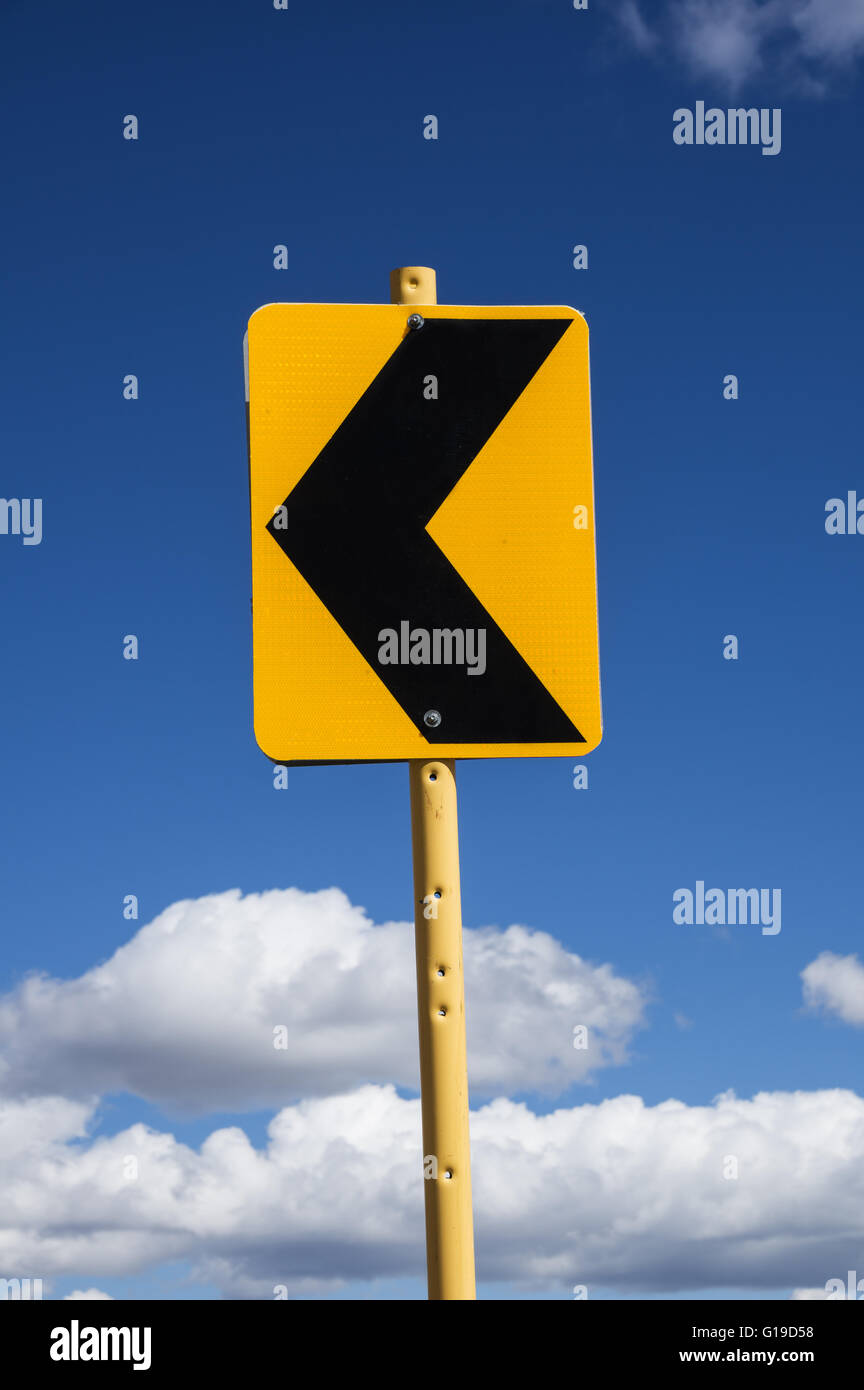 sign with black arrow pointing left warning of a curving road with blue sky and clouds background Stock Photo