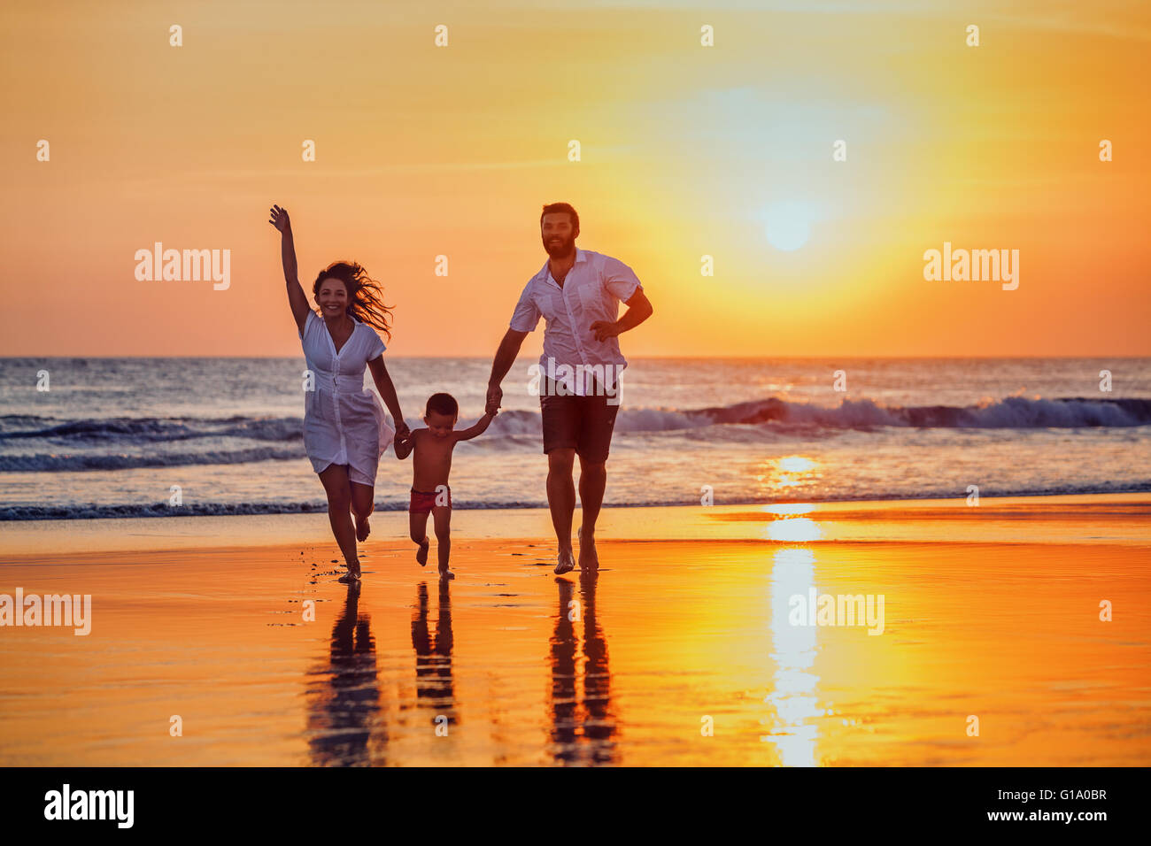 Happy family - father, mother, baby son hold hands and run with fun along sunset sea surf on black sand beach. Stock Photo