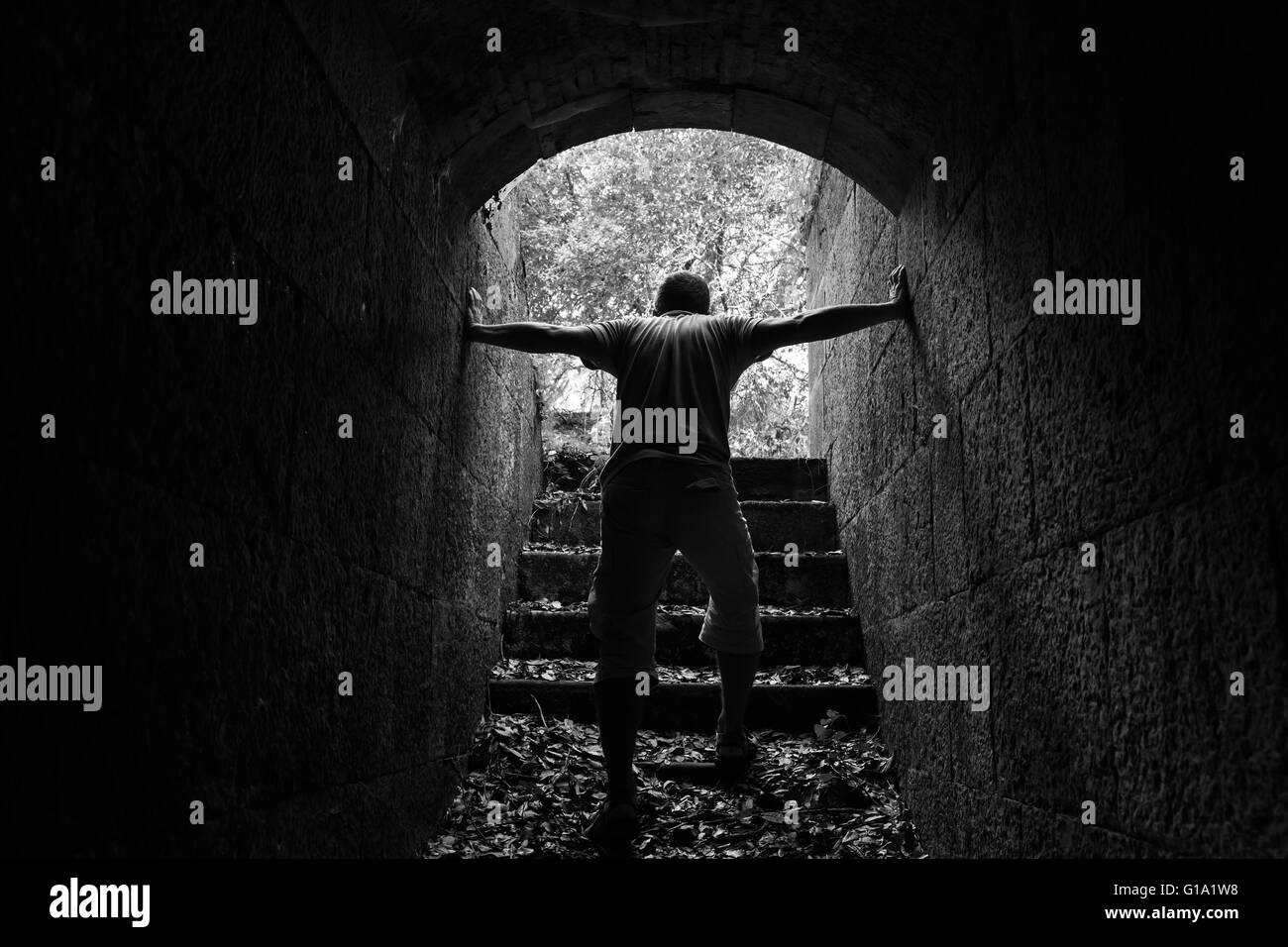 Tired man goes out of dark stone tunnel with glowing end, monochrome photo Stock Photo