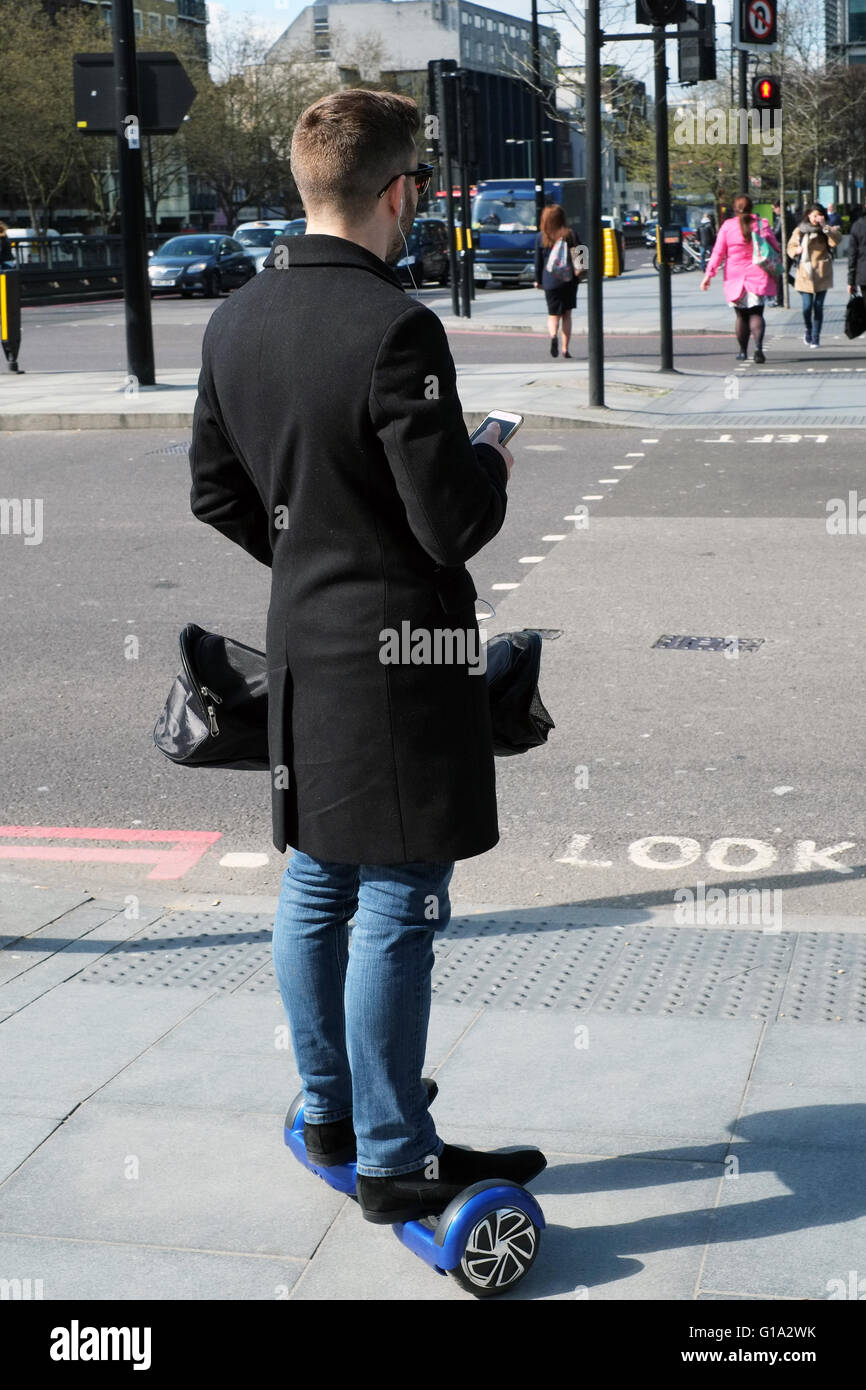 A young man checks his smartphone as he balances on an electric powered scooter board on the Euston Road, in London, England, UK Stock Photo
