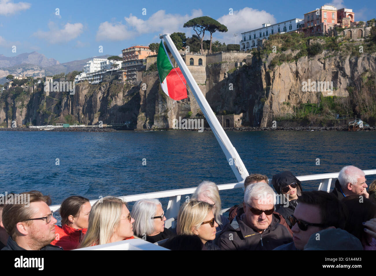 Tourists sitting in a ferry boat with an Italian flag returning from Capri to Sorrento in the Gulf of Naples with Sorrento's cliffs behind. Italy Stock Photo