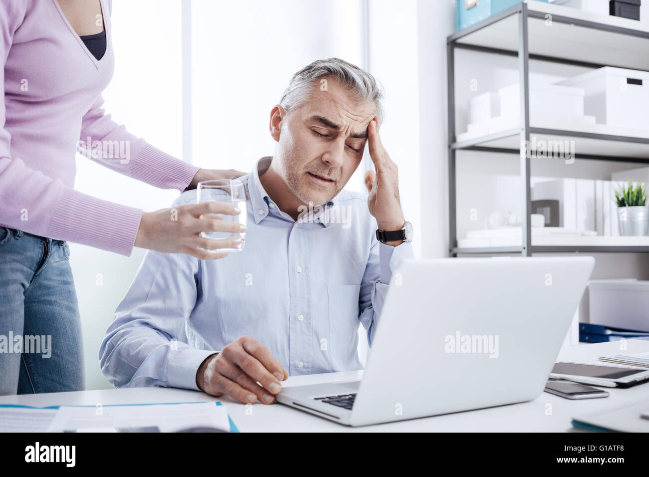 Tired businessman in the office working at desk and having a bad headache, his colleague is giving him a glass of water and touc Stock Photo