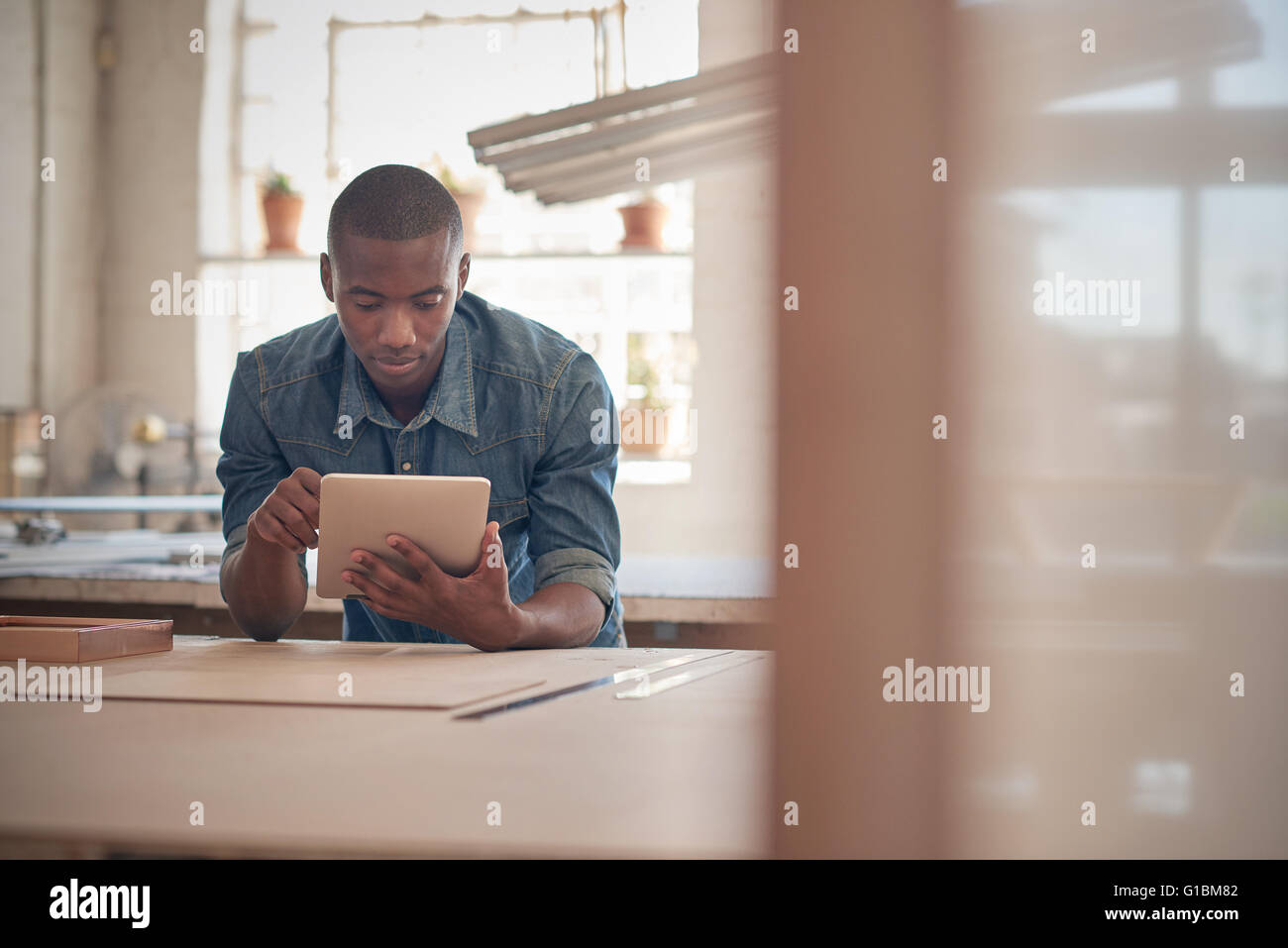 Handsome young entrpereneur of African descent, leaning on a work table in a design studio, using a digital tablet Stock Photo