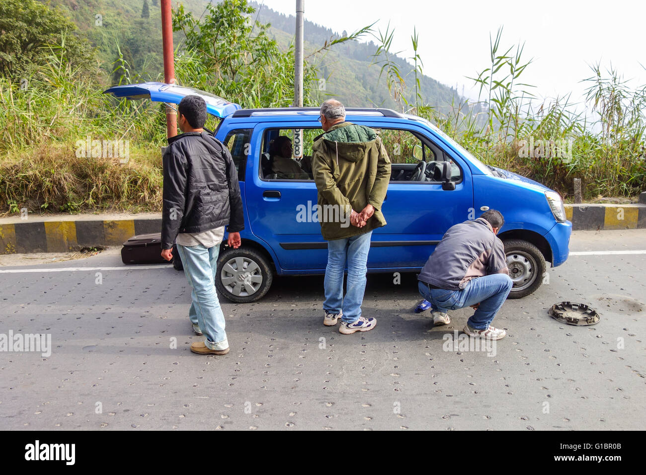 A private taxi owner replaces the punctured tire of his Wagon-R car while the stranded passengers wander on National Highway 55 Stock Photo