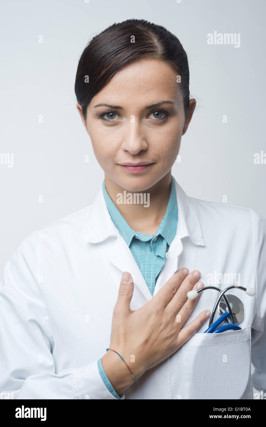 Attractive female cardiologist with stethoscope and lab coat, touching her chest. Stock Photo
