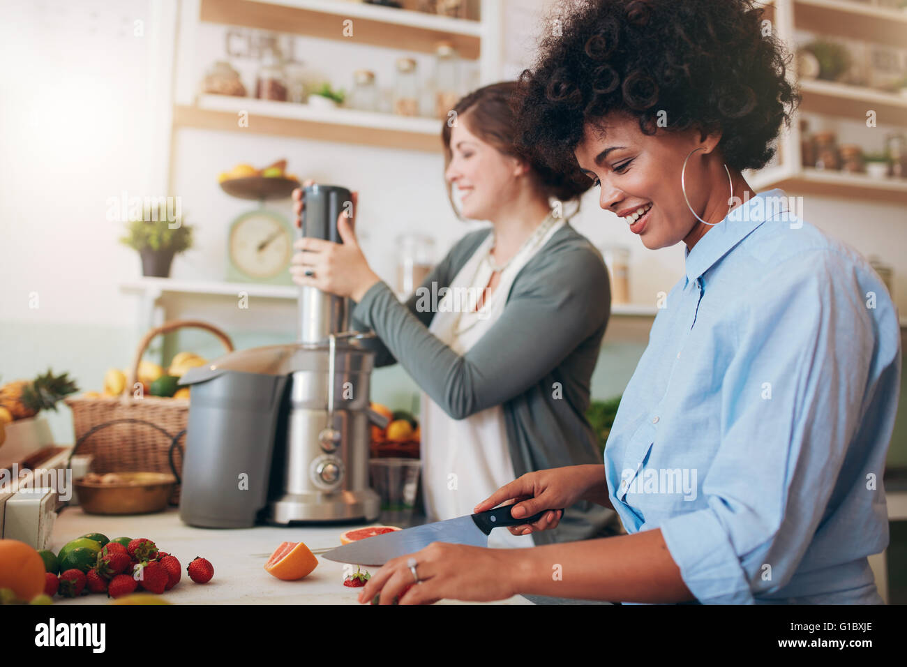 Smiling young woman cutting fruits with coworker using juicer in background, two women preparing fruit juice at cafe counter. Stock Photo