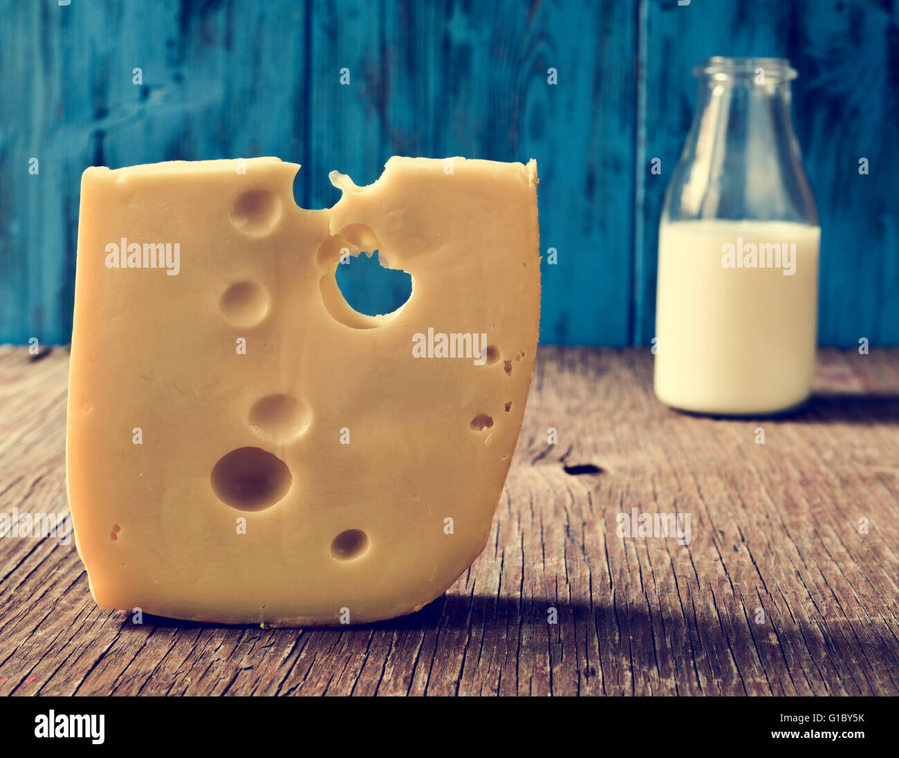 closeup of a piece of Swiss cheese and a glass bottle with milk on a rustic wooden table, against a blue rustic wooden backgroun Stock Photo