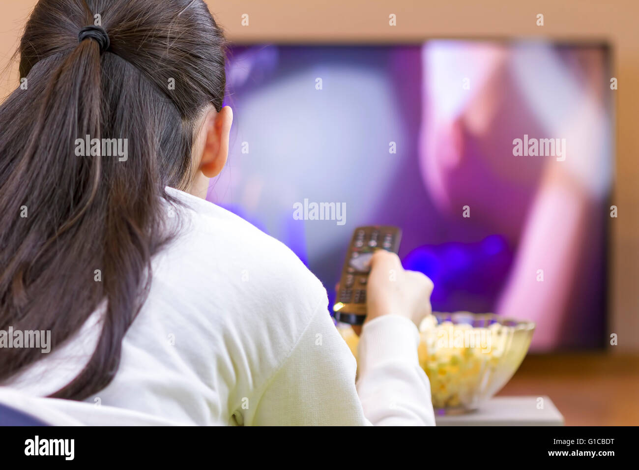 Teenager girl with remote control laying down and watching smart tv eating popcorn Stock Photo