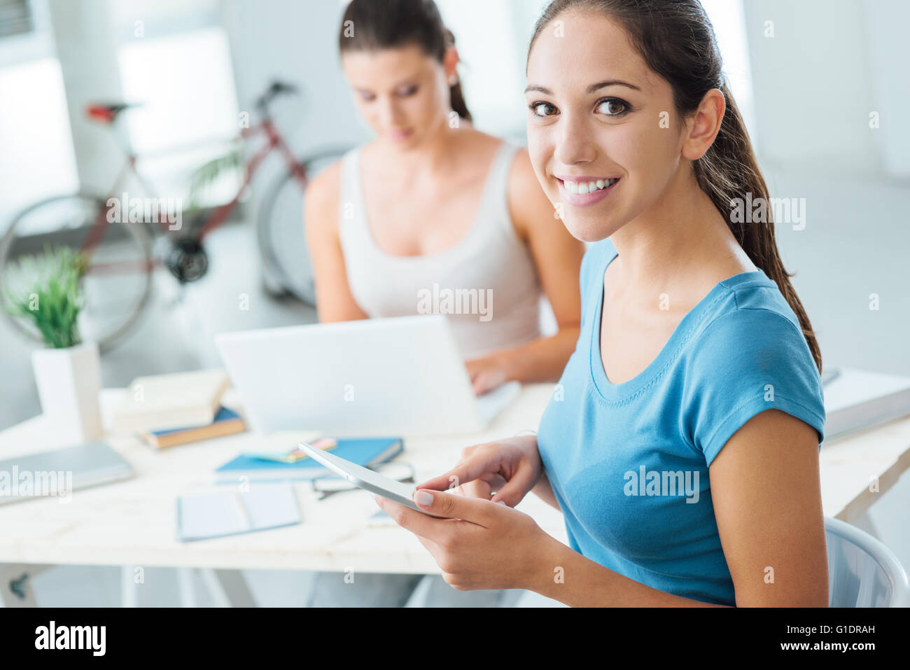 Cute young girl using a digital touch screen tablet and smiling at camera, her friend is sitting at desk and using a laptop Stock Photo