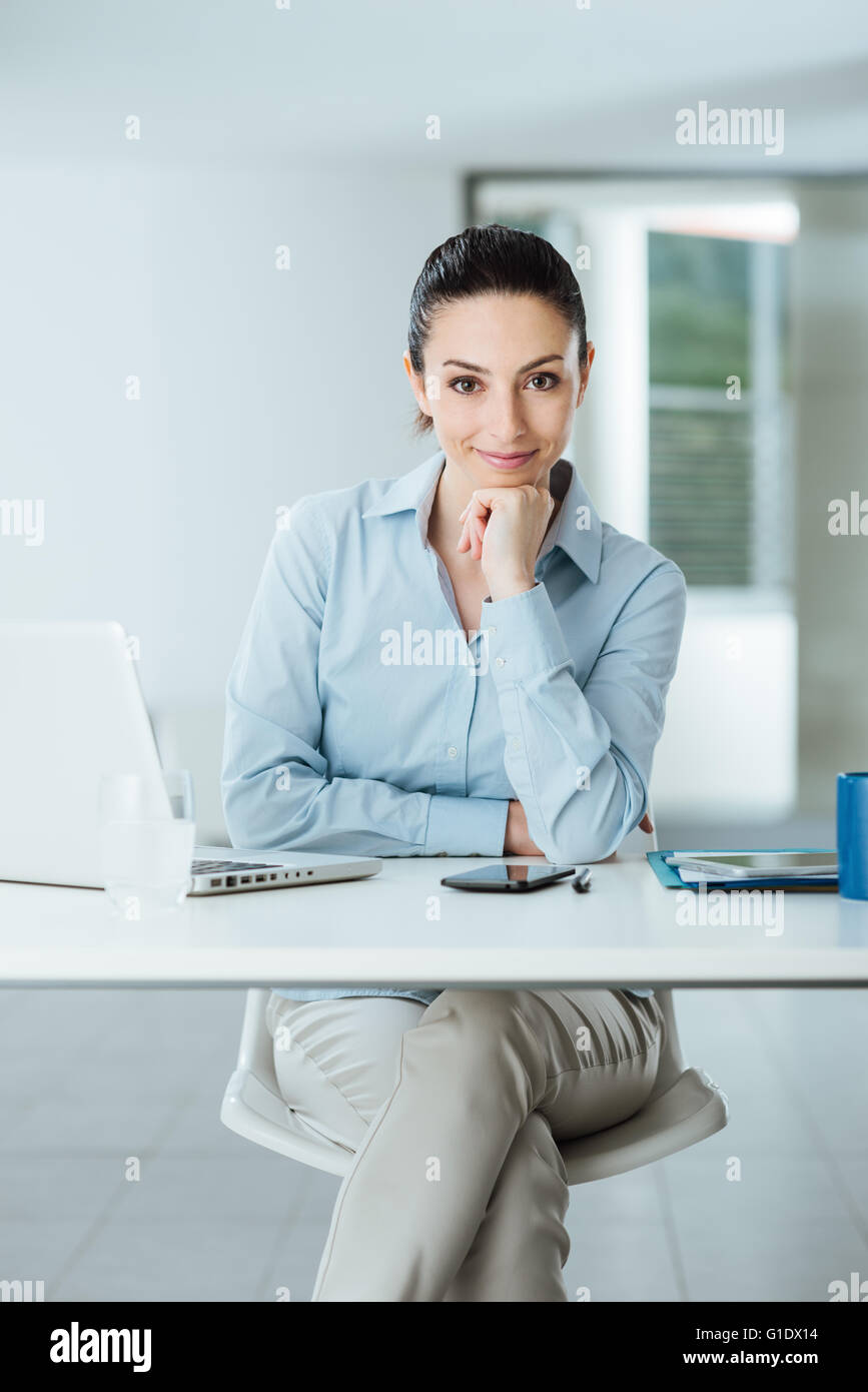 Beautiful confident female manager sitting at office desk and smiling at camera with hand on chin, room interior on background Stock Photo