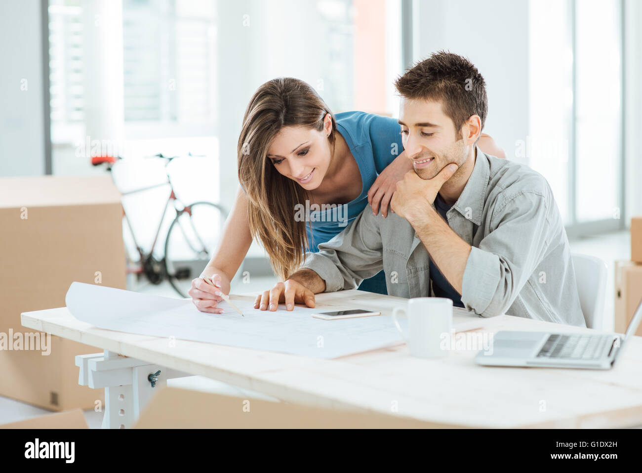 Yougn couple planning their new dream house, he is sitting at desk and she is drawing on a project, carton boxes on background Stock Photo