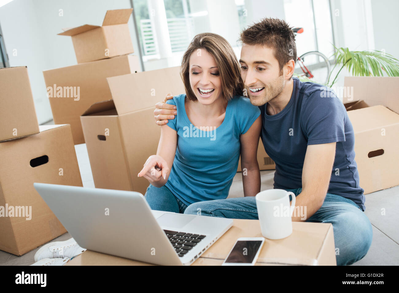 Smiling couple sitting on their new house floor surrounded by carton boxes, they are watching a funny video on a laptop and laug Stock Photo