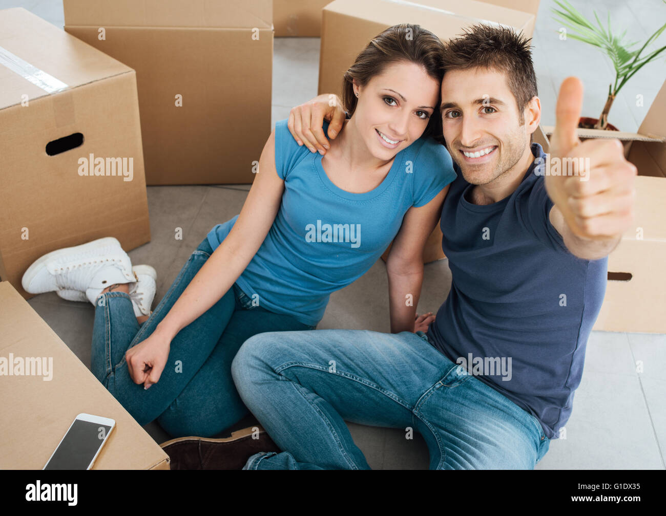 Happy young couple thumbs up and smiling at camera, they are sitting on their new house floor surrounded by carton boxes Stock Photo