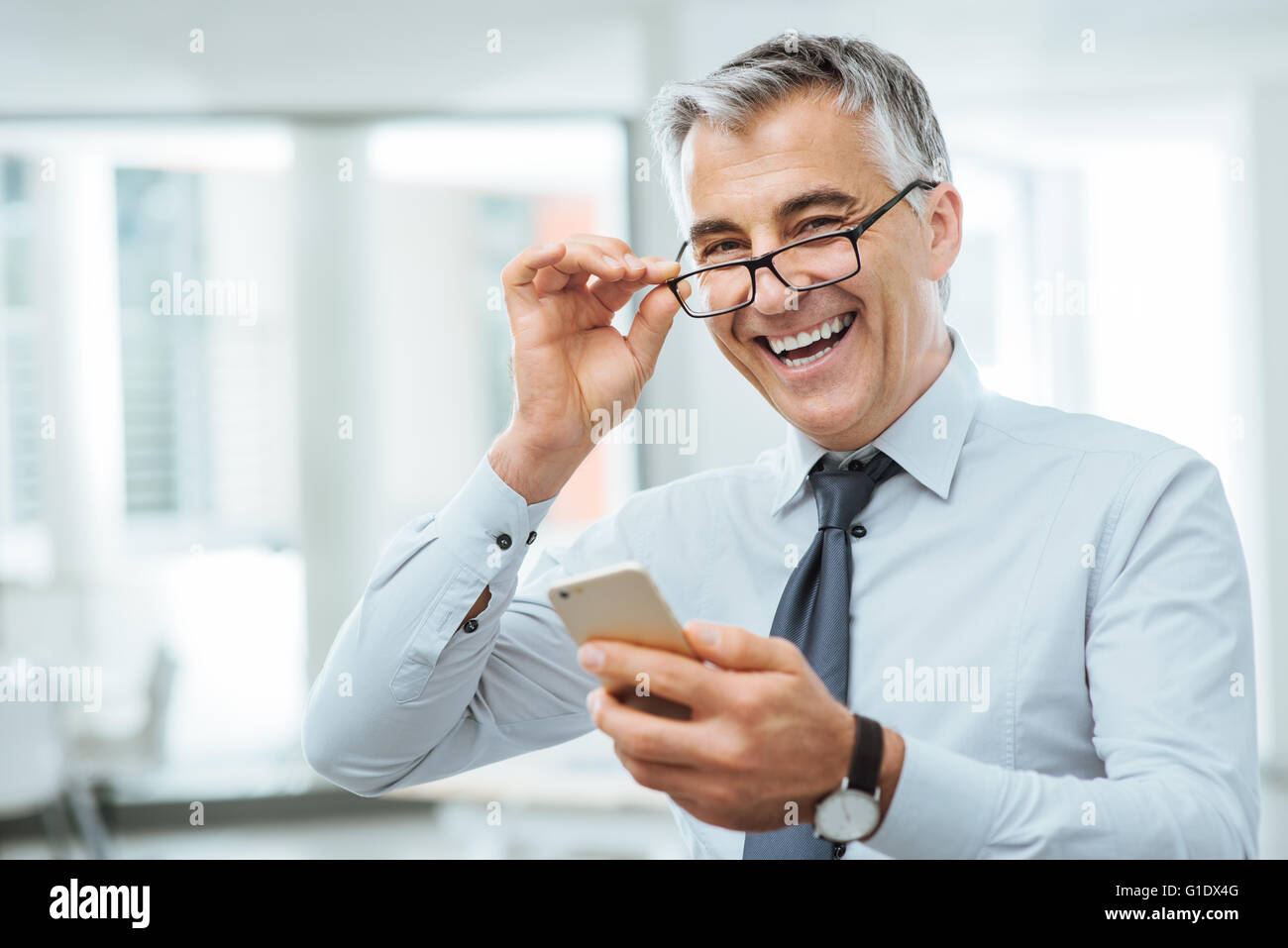 Smiling businessman with eyesight problems, he is adjusting his glasses and reading something on his mobile phone Stock Photo