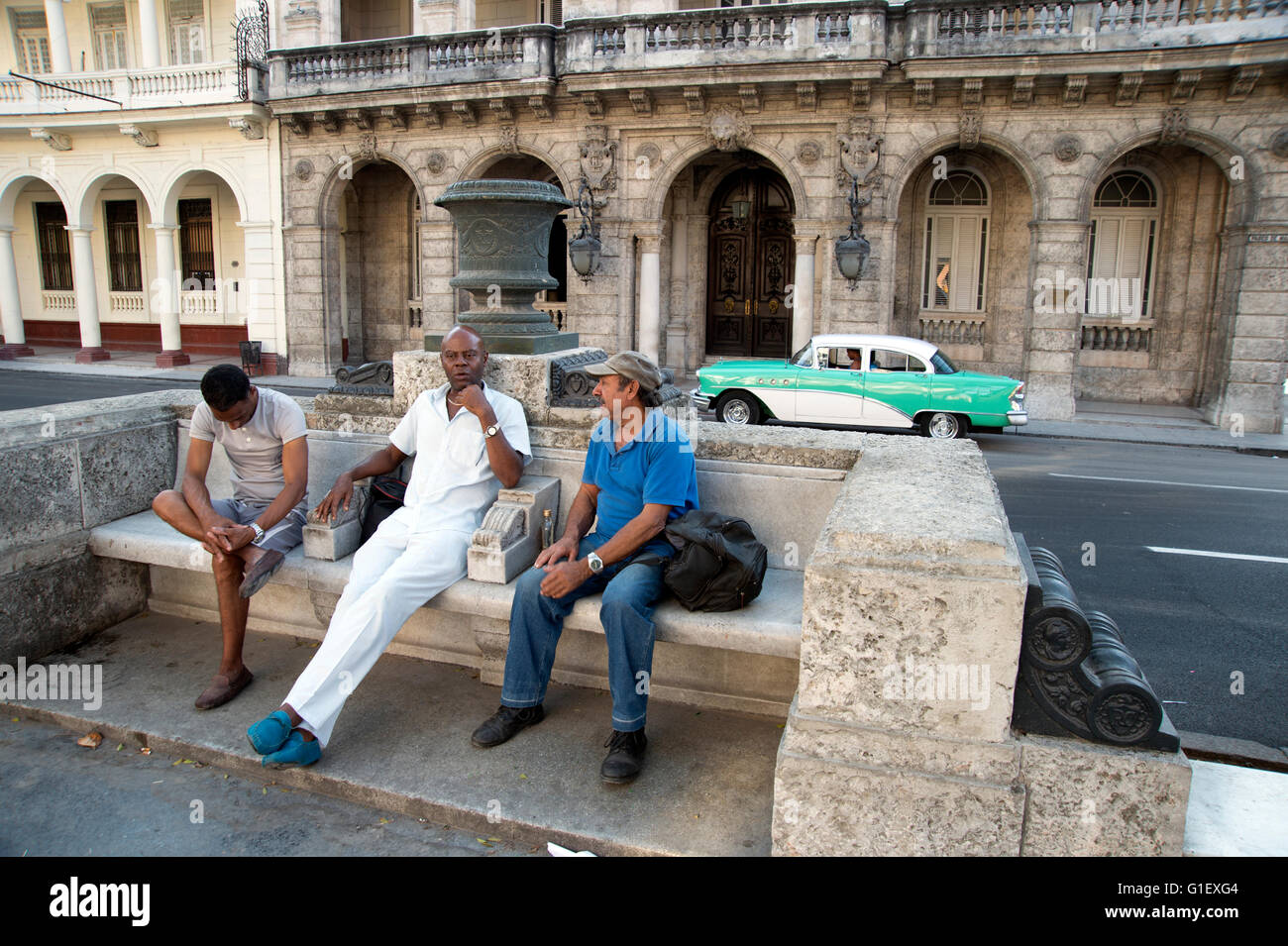 Three Cuban men sit on a stone bench on the Prado in Centro Havana Cuba with an 1950's American car in the background Stock Photo