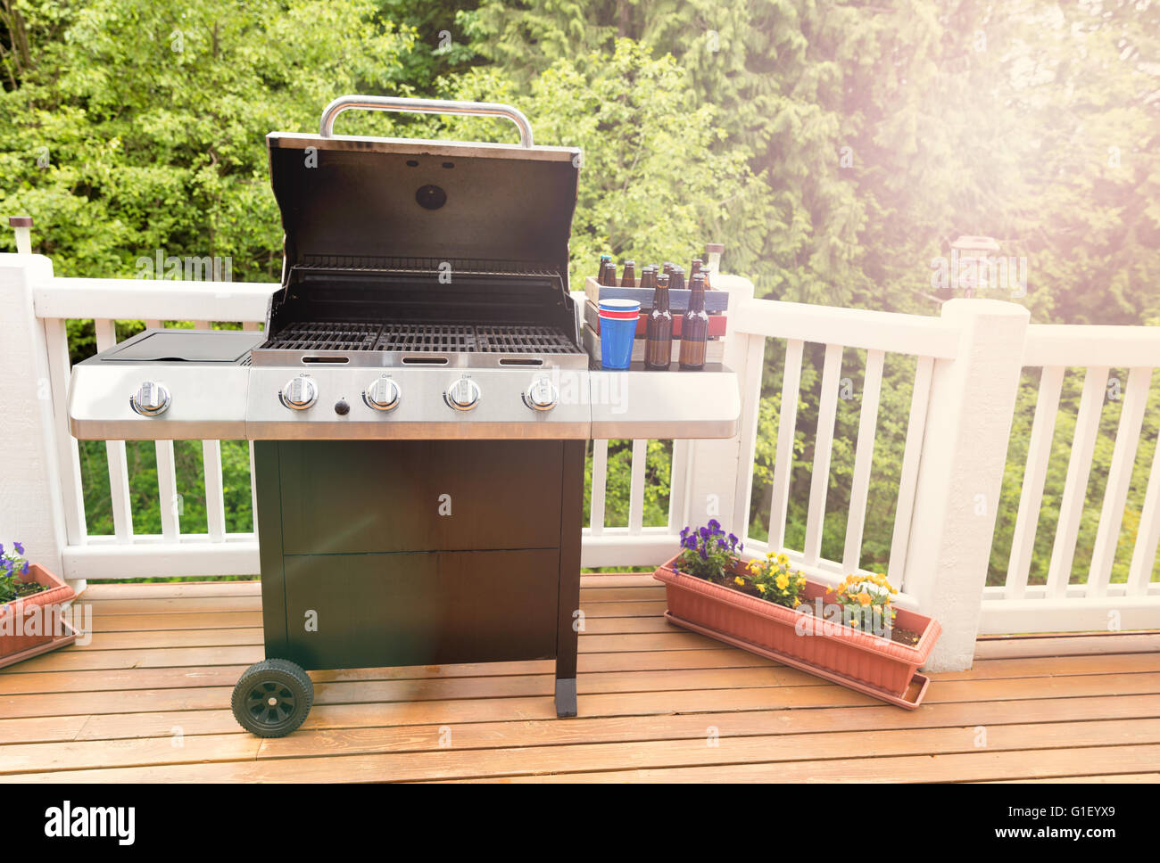 Bright daylight falling on open large barbecue cooker, bottled beer, cup, and crate on cedar wood deck with trees in background. Stock Photo