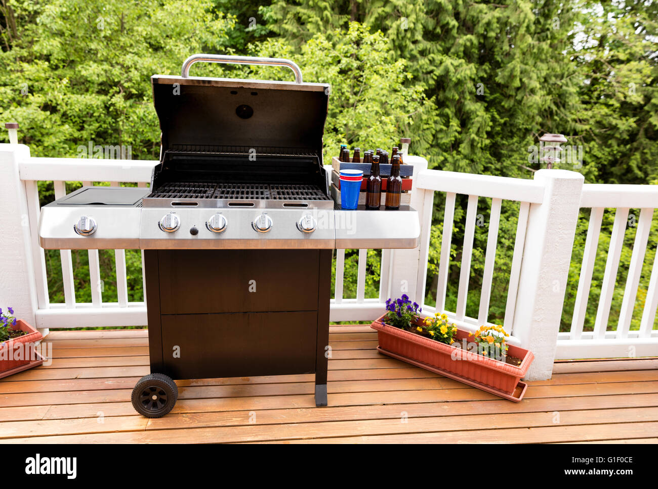 Open large barbecue cooker, bottled beer, cup, and crate on cedar wood deck with trees in background. Stock Photo