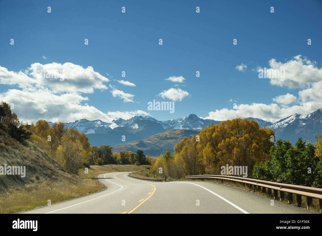 a winding mountain road goes between yellow aspen trees towards distant mountains Stock Photo