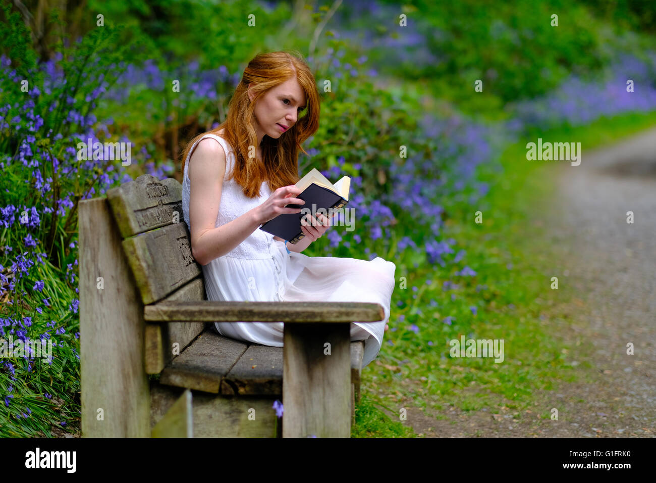 A young woman with long red hair and in a white dress sits reading a ...