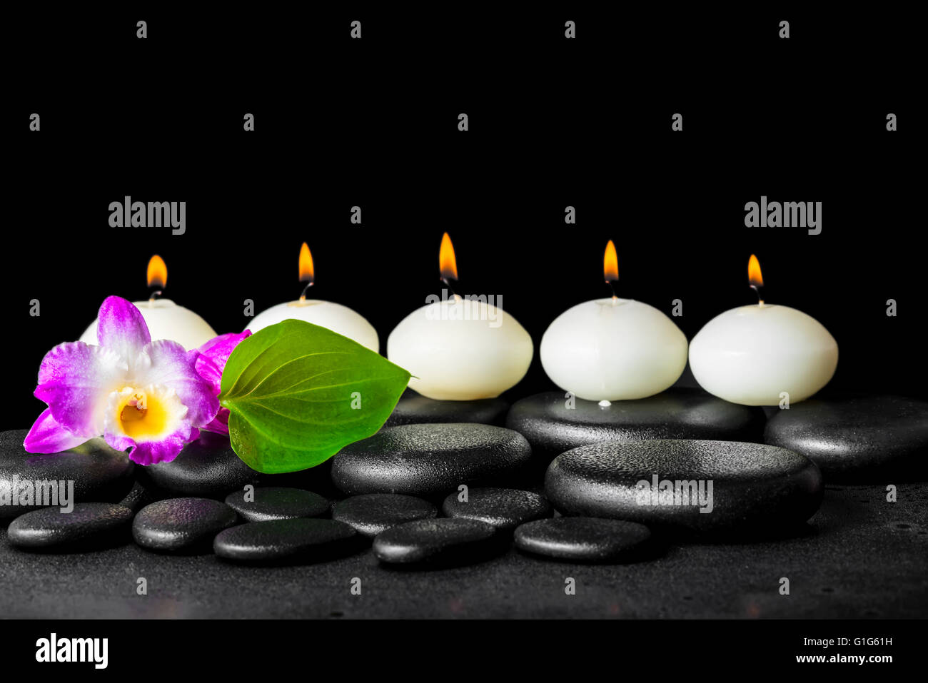 spa still life of row white candles, orchid flower dendrobium and green leaf on black zen stones background with dew, closeup Stock Photo