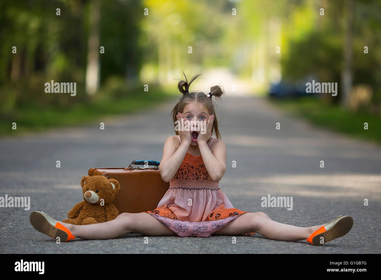 Funny little girl with a suitcase and teddy bear sitting on the road. Stock Photo