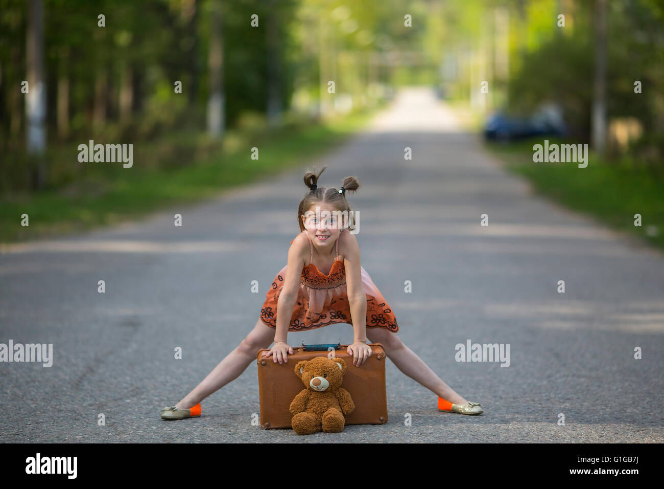 Funny little girl with a suitcase and teddy bear on the road. Stock Photo