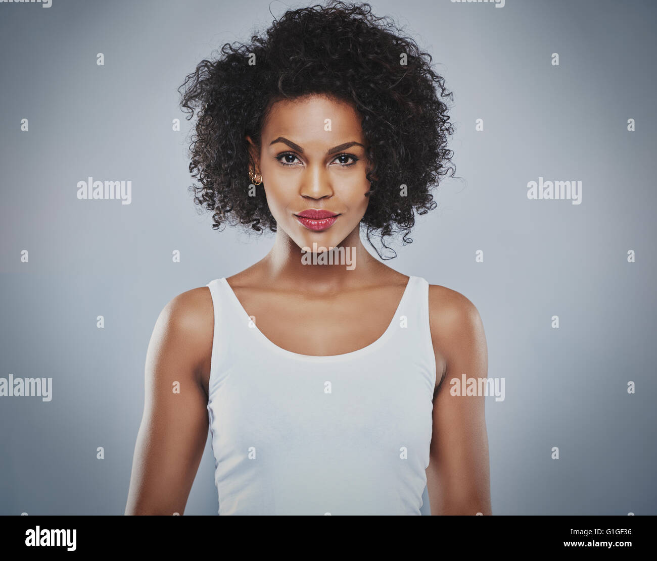 Centered front view of single pretty grinning woman wearing white sleeveless undershirt and calm expression over gray background Stock Photo