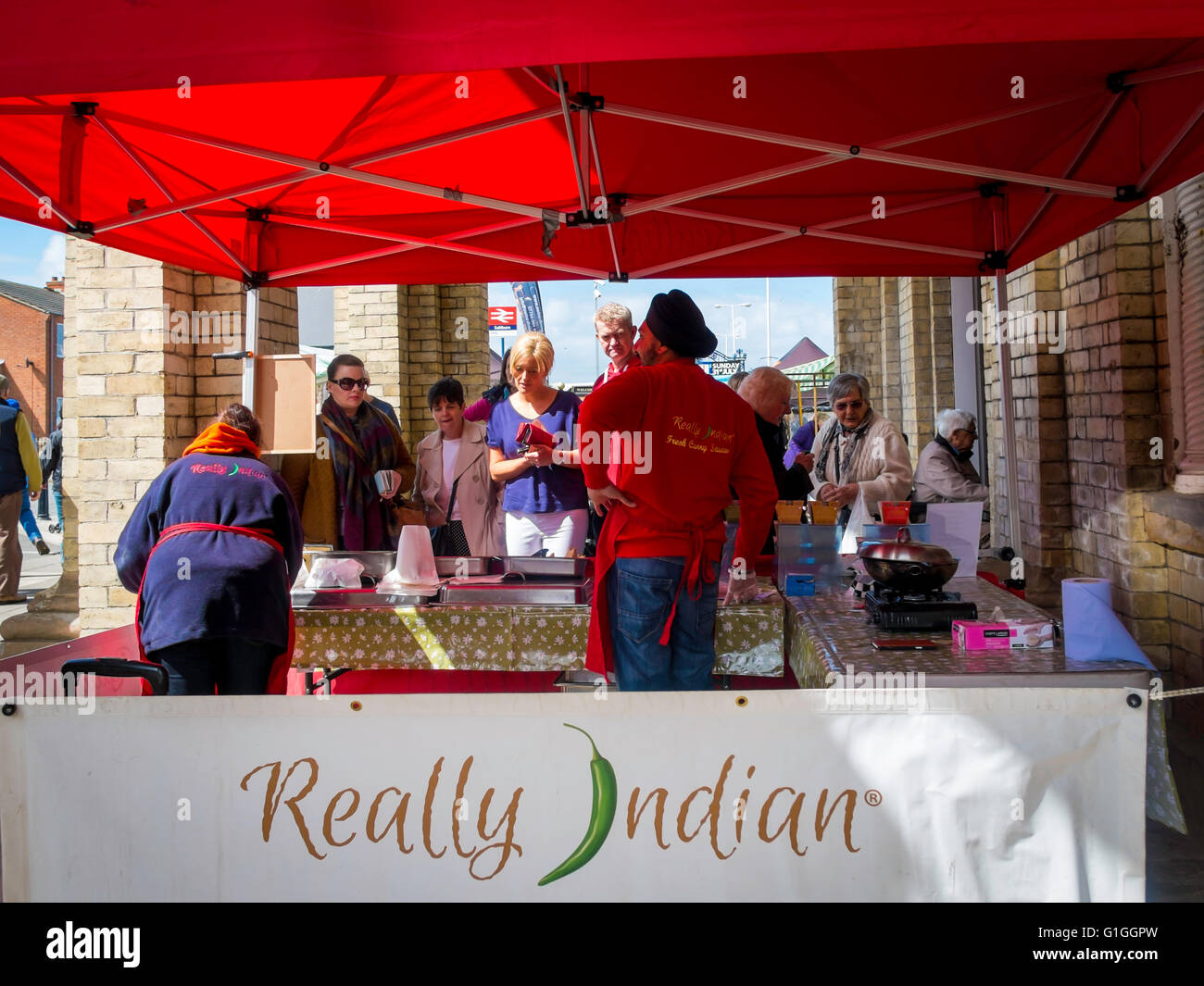 Sikh man and woman stallholders at a UK farmer's market serving cooked 'Really Indian' food to a crowd of people waiting to buy Stock Photo