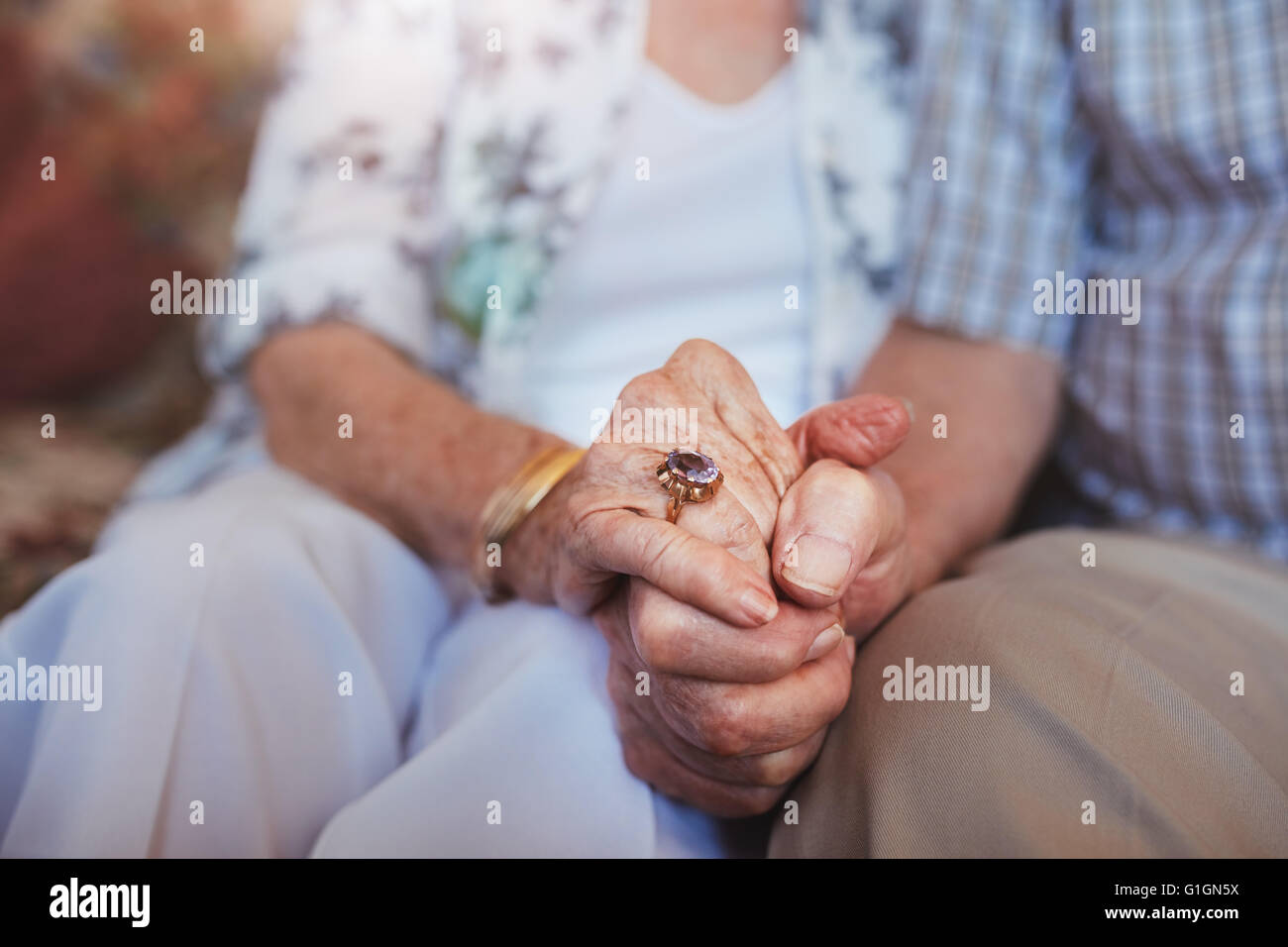 Cropped shot of elderly couple holding hands while sitting together at home. Focus on hands. Stock Photo