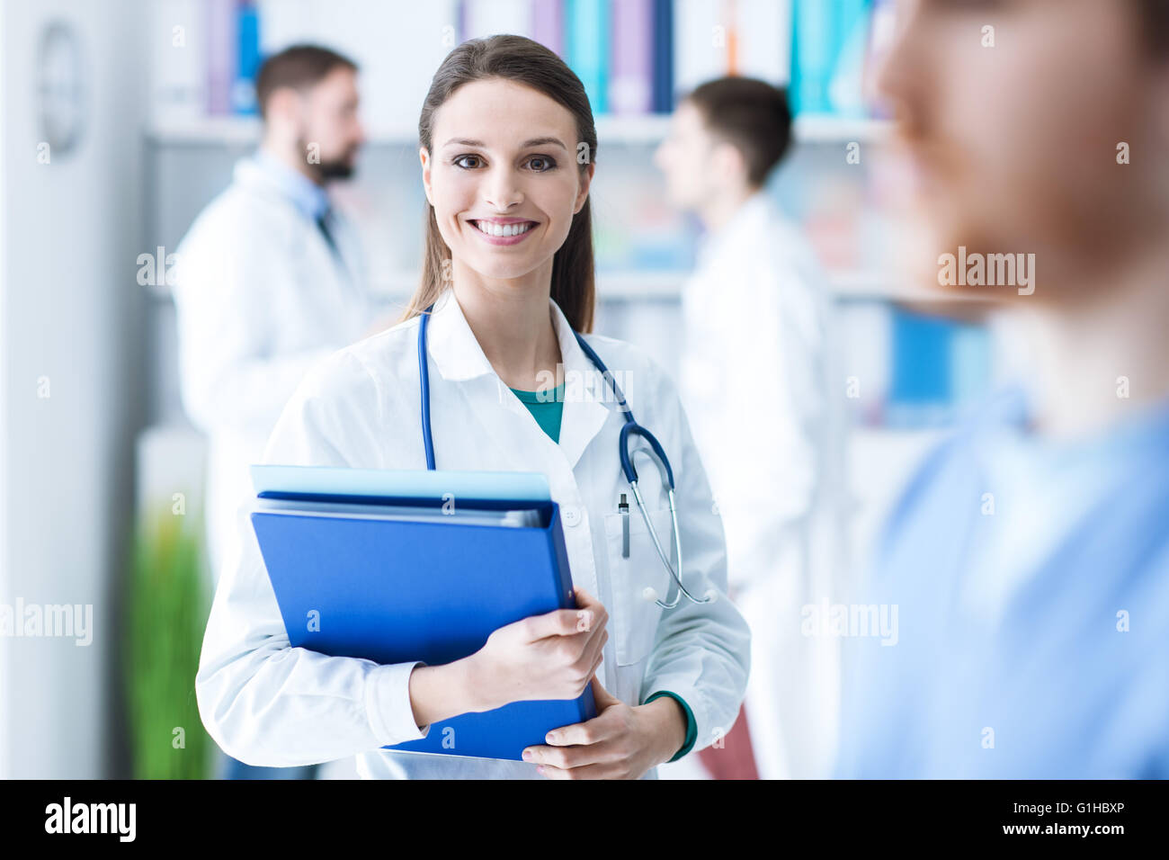Confident attractive female doctor in the office holding medical records and folders, she is smiling at camera, medical staff on Stock Photo