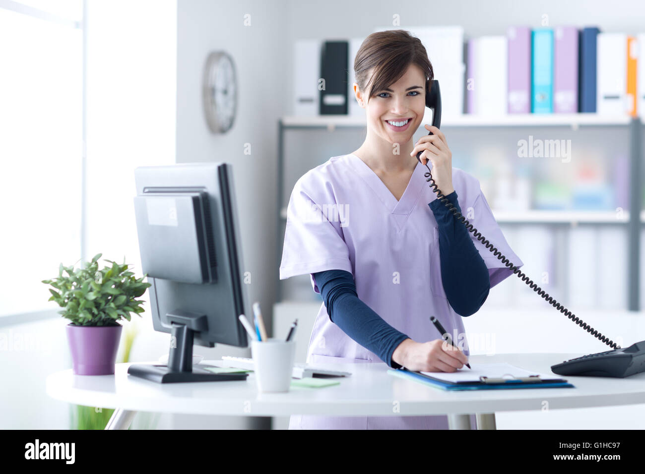 Young practitioner doctor working at the clinic reception desk, she is answering phone calls and scheduling appointments Stock Photo