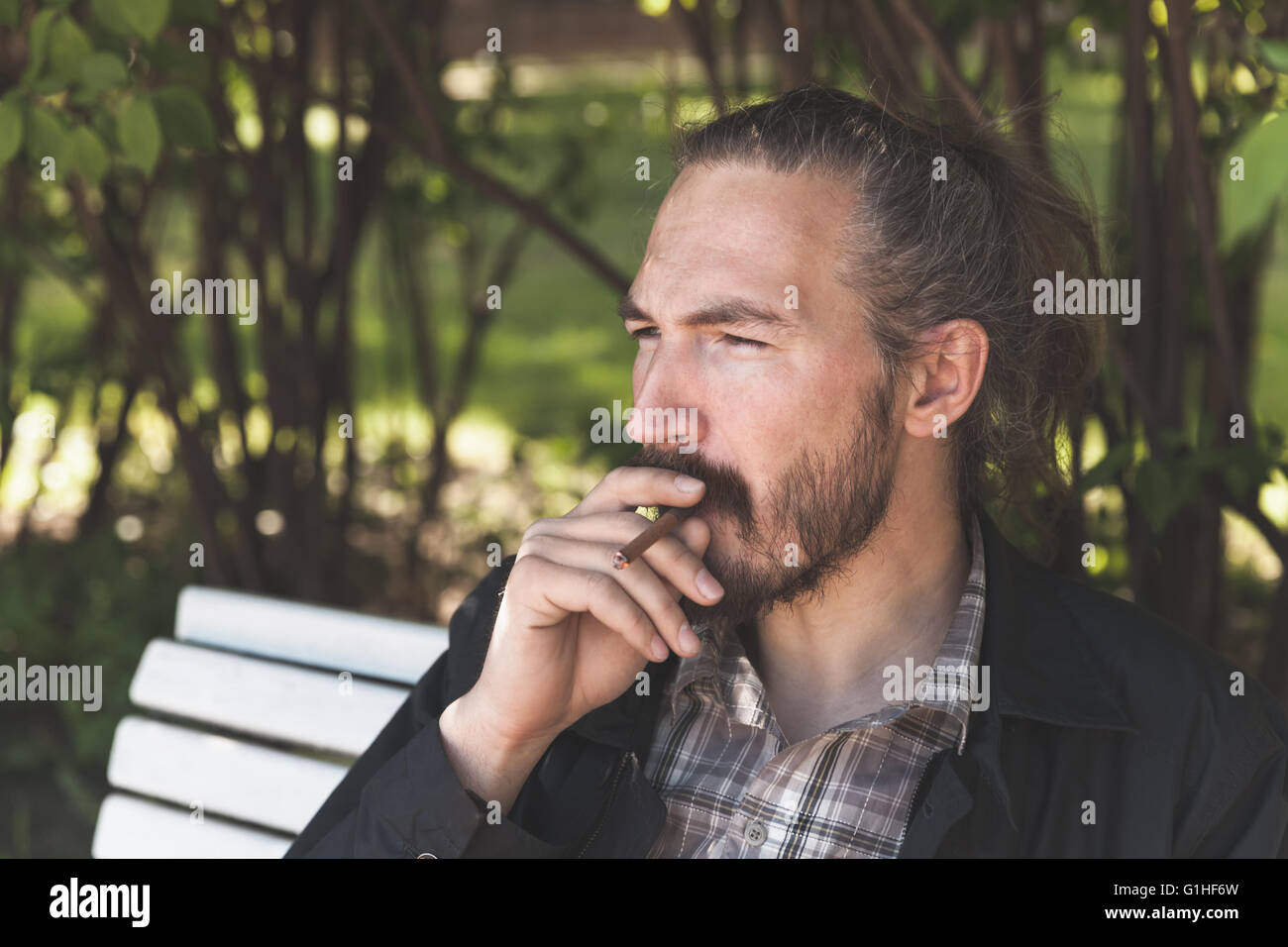 Bearded Asian man smoking cigar in summer park, outdoor portrait with selective focus Stock Photo