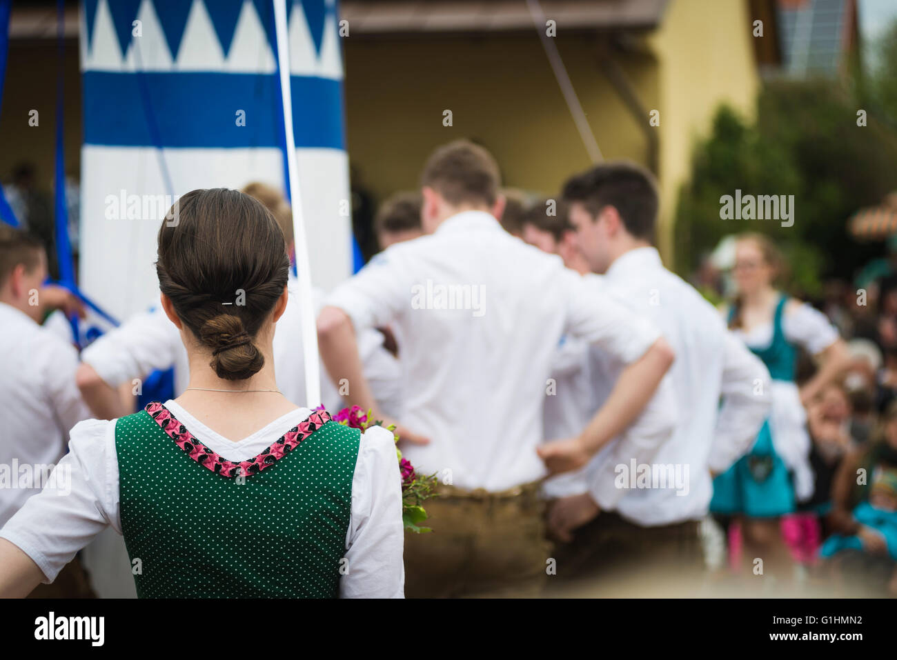 Woman in local dress watching young men in Lederhose standing around a maypole while dancing a traditional Bavarian folk dance Stock Photo