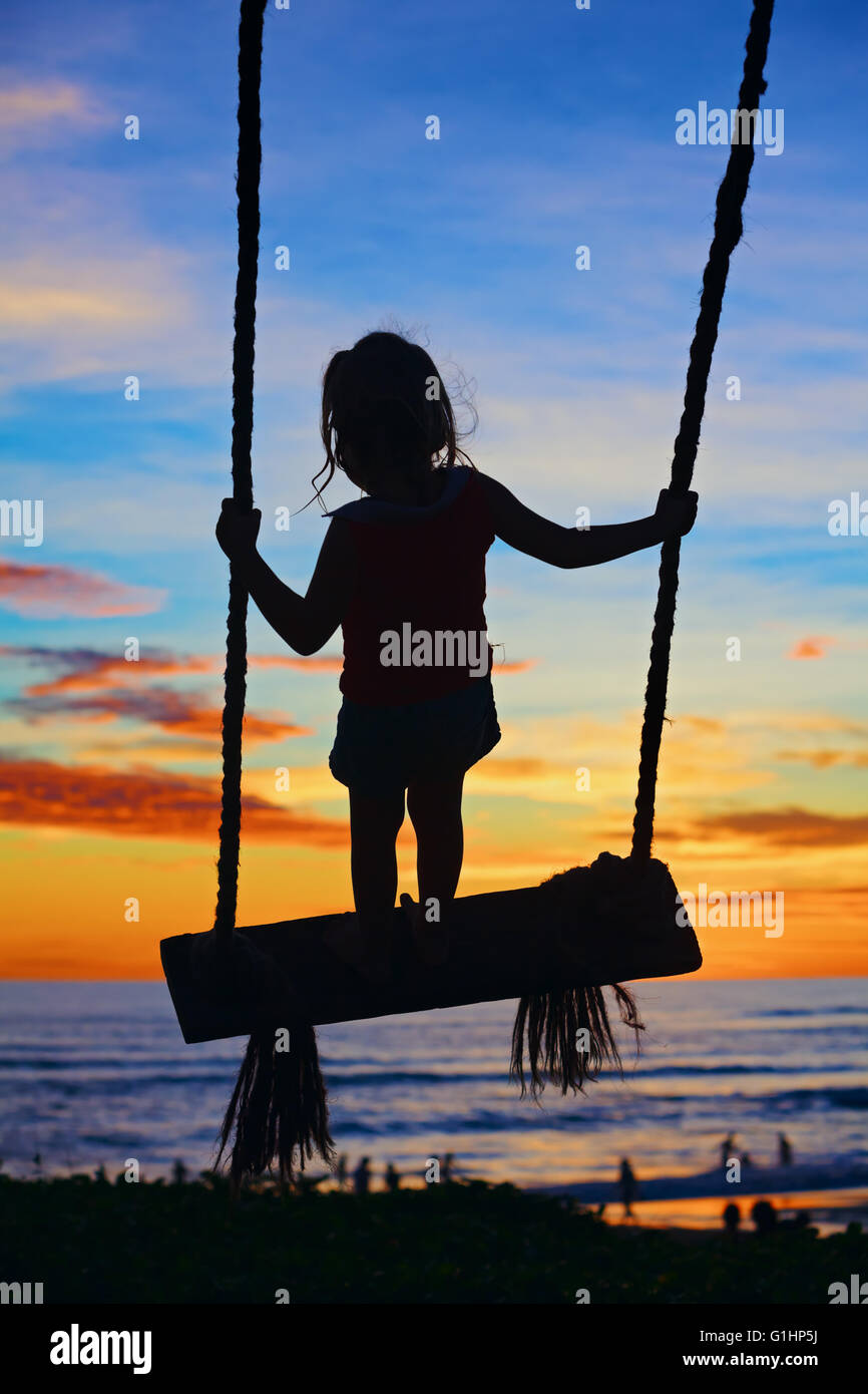 Black silhouette of happy baby girl flying high with fun on swing on ocean beach surf and blue orange sunset sky background. Stock Photo