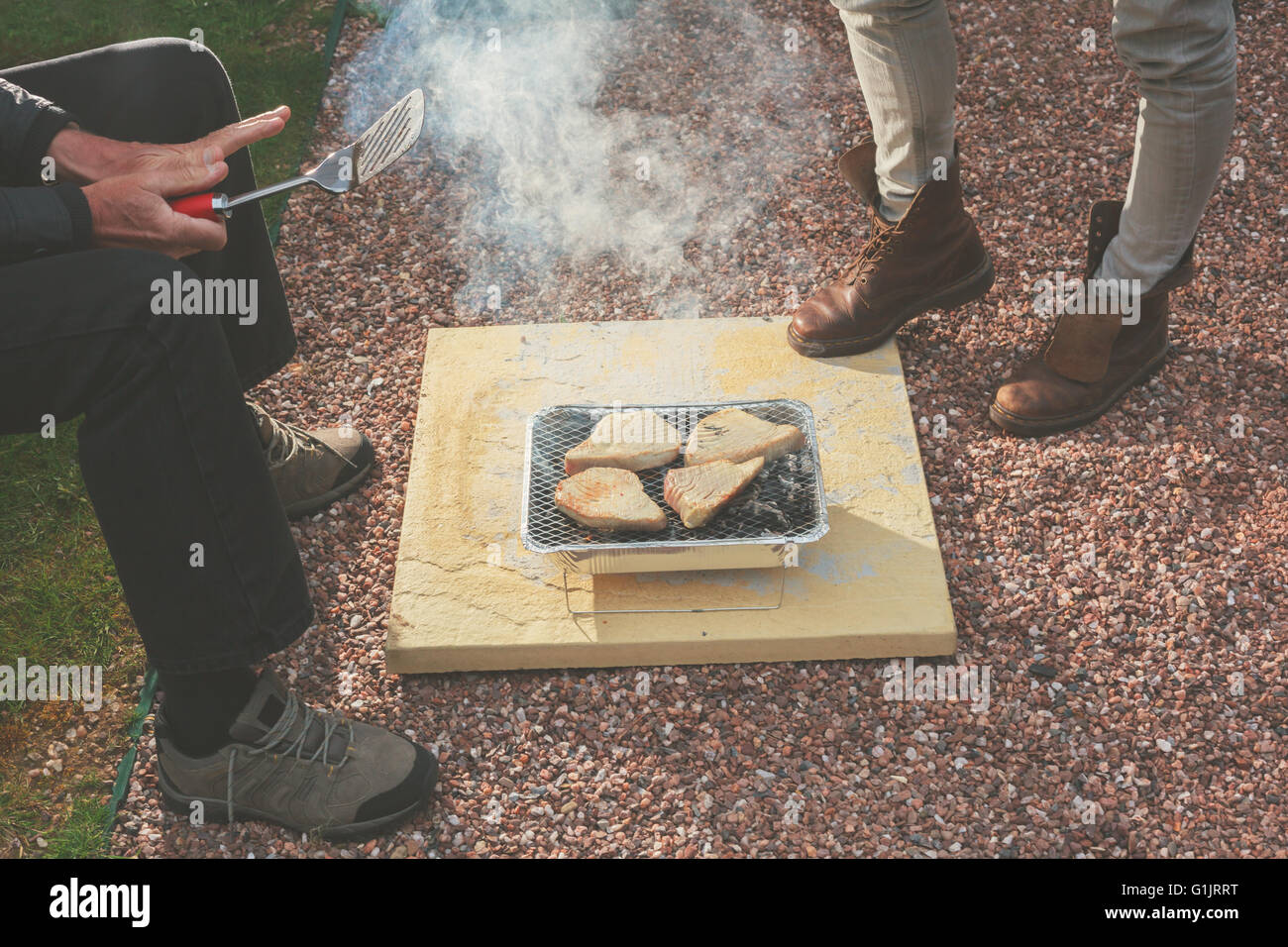 Two people are standing around a disposable barbecue outside Stock