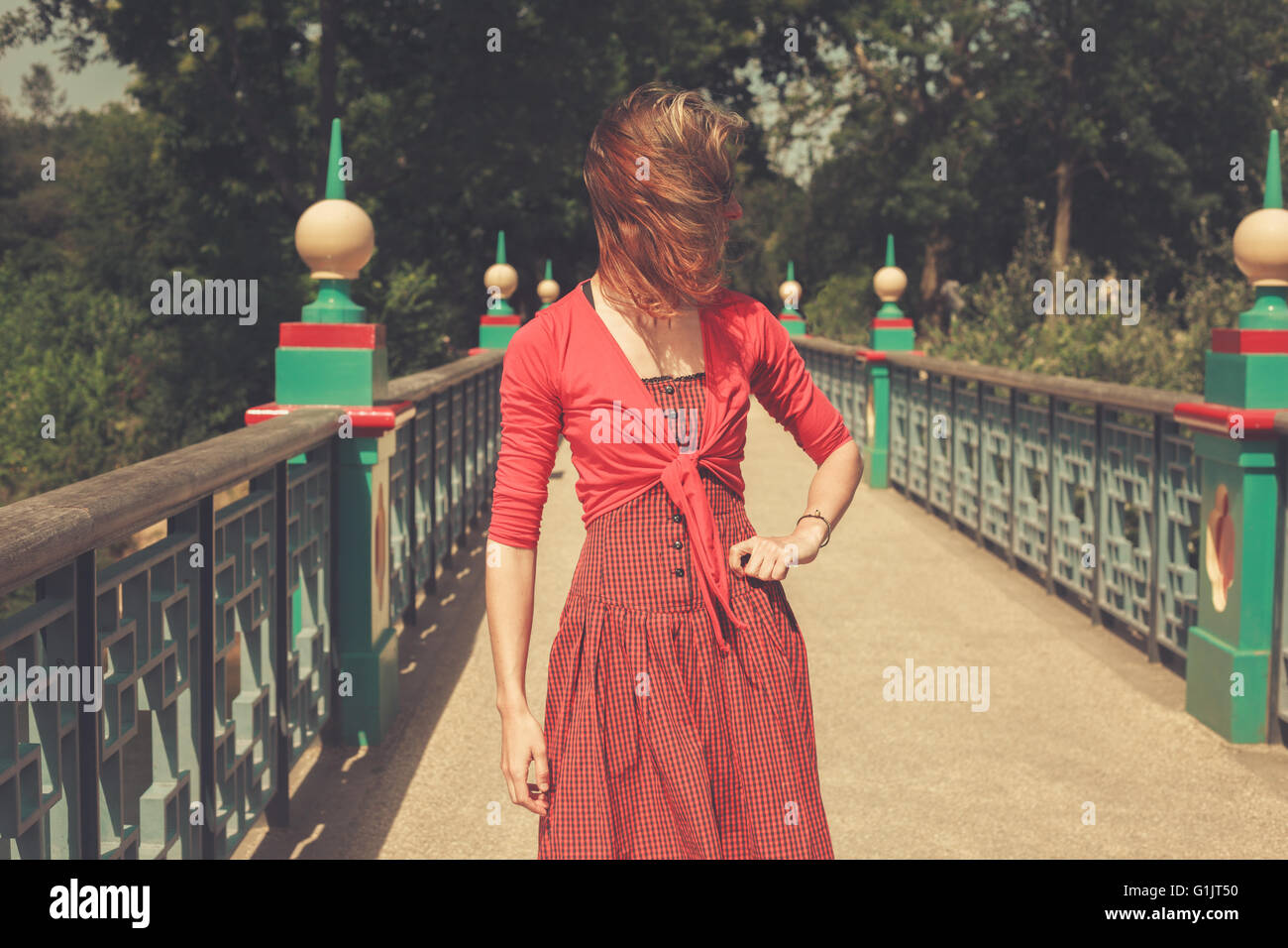 A young woman wearing a red dress is standing on a bridge in a park ...