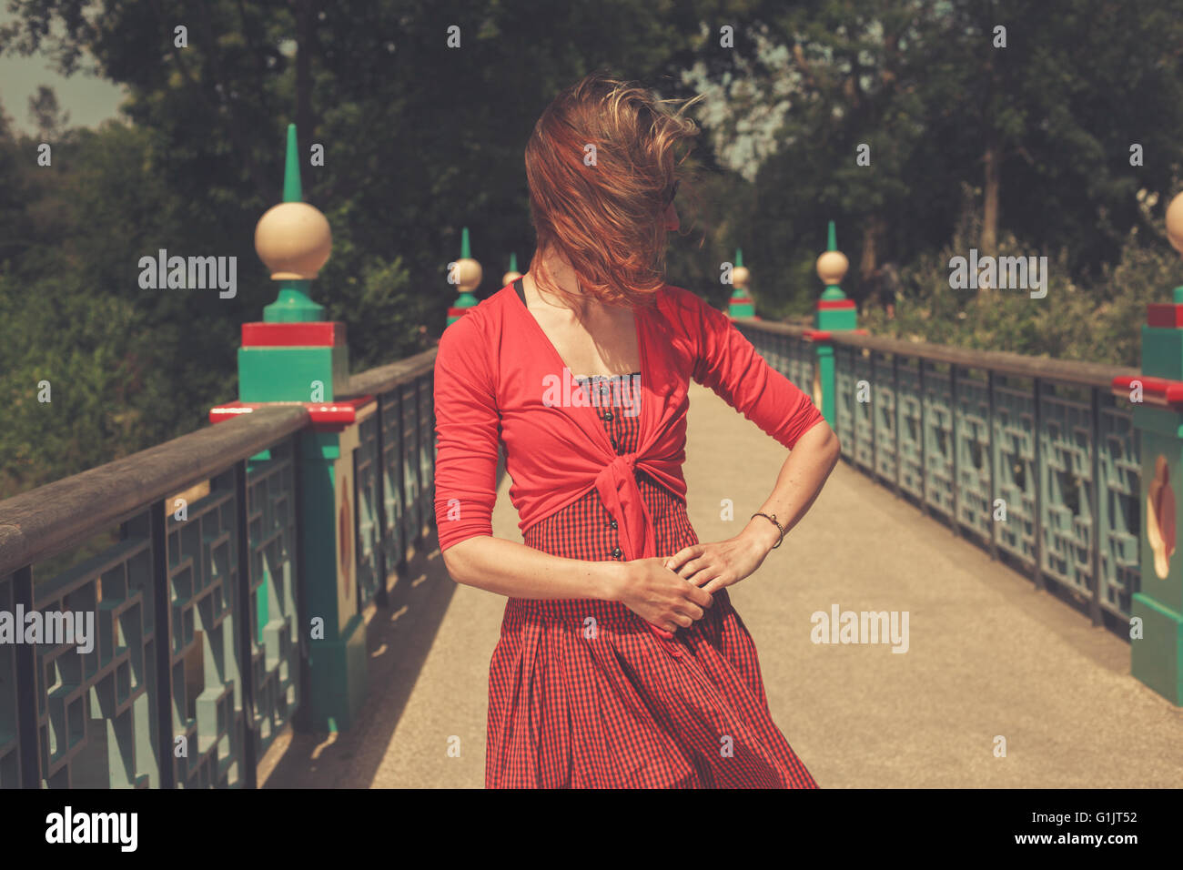 A young woman wearing a red dress is standing on a bridge in a park ...