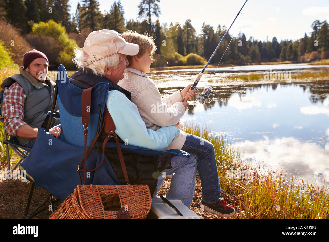 Grandad teaches his grandson to fish at a lake, dad watching Stock Photo